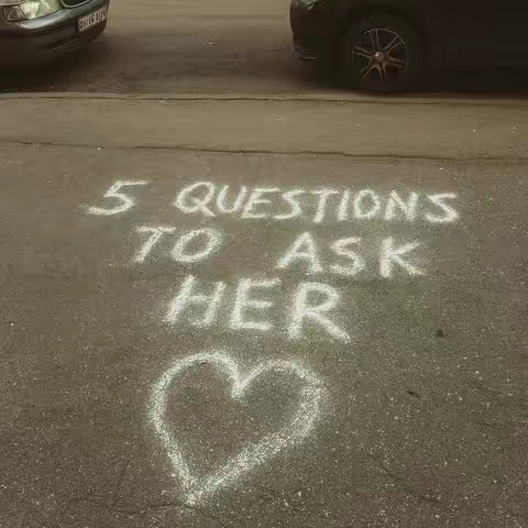 Street-level shot featuring text written on the asphalt. Two cars, one on each side, frame the scene. In the center, large text says '5 QUESTIONS TO ASK HER' in chalk-like font. Below it is a heart symbol, completing the emotional message.