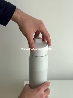 A person is opening a white water bottle; the background is a plain wall, and the person's hands and part of their arm are visible, wearing a black sleeve.