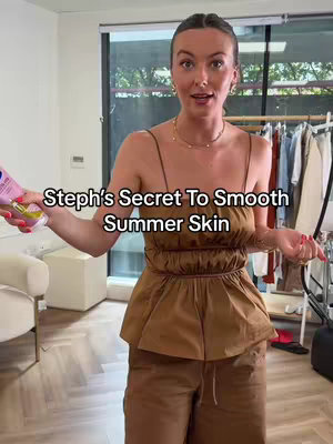 A woman in a brown top and pants is holding a tube of lotion to the left, standing in front of an open window with blinds, a white stand, and a clothing rack with clothes