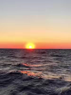 A serene seascape at sunset. The sun, a bright orb of yellow, is positioned near the horizon, casting a warm glow across the water. The sky transitions from a light blue at the top to a rich orange and red near the horizon. The ocean's surface is subtly detailed with gentle waves, reflecting the light and colors of the sky. Overall, the composition is peaceful and picturesque.