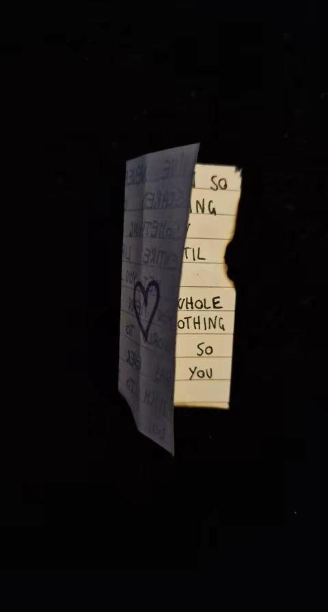 A partially torn piece of paper with handwritten text is positioned against a black background. The paper, likely white or cream-colored, reveals snippets of phrases. A heart is drawn on the left side of the paper. The right side shows the text, illuminated by a bright light. The backdrop is completely black.