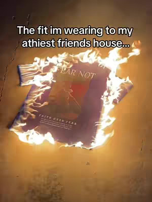 A purple T-shirt with gold text and the phrase "FEAR NOT" is engulfed in flames against a brown background. The text "FAITH OVER FEAR" is visible on the bottom of the shirt. White flames lick around the edges of the shirt.