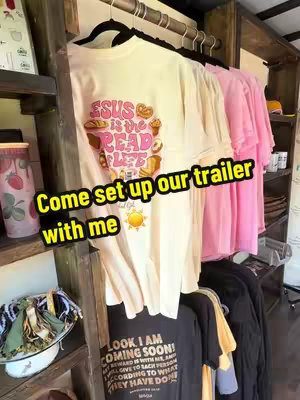 The image shows an interior view of a store or trailer. Clothes are hanging on racks. A t-shirt with the words "JESUS IS THE BREAD OF LIFE" is visible. Additional text overlays the image, and the setting has a rustic design with wooden elements.