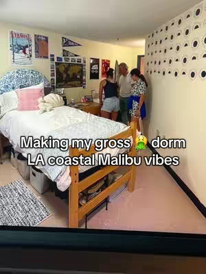 A dorm room setup featuring a bed on the left, with bedding and posters above. There are three people visible in the background, and a text overlay that reads "Making my gross 🤢 dorm LA coastal Malibu vibes". The room’s color scheme is muted.