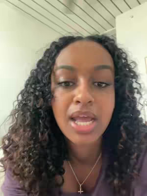 A woman with curly black hair looks directly at the camera speaking. She is wearing a purple shirt and a silver necklace with a cross pendant. The background is a simple white wall and ceiling.