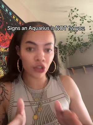 A woman with dark hair wearing a white tank top and gold hoop earrings is speaking to the camera. A colorful tapestry hangs in the background, along with some leafy green plants.