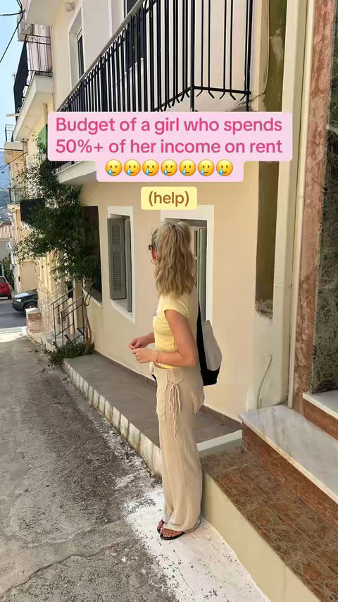 A woman with blonde hair is seen from the back, standing in a narrow street. She is facing away from the camera and looking towards the right. She is wearing a yellow shirt and light-colored pants. The background includes a building, a street with other buildings and a sign.