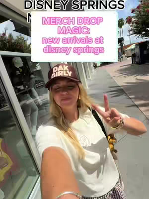 A selfie-style shot taken at Disney Springs, Florida. The creator, a woman with blonde hair and a "Dak Girly" hat, is in the foreground, making a peace sign. Behind her is a storefront with a large window, and in the background is a walkway with other shops or buildings. A text overlay reads "Merch Drop Magic: new arrivals at disney springs."