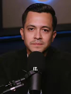 A man with dark hair and a black jacket is centered in the frame. He's looking at the camera with a neutral expression. A black microphone obscures part of his lower face. The background is a blurry mix of dark colors with some blue lighting. The text 'IT.' is written on the cover.