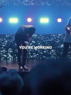 The image shows a figure on a stage, with a lowered head and hands clasped as if praying. The background is a blurry, colorful, stage lighting setup. Text overlayed on the image reads 'YOU'RE WORKING'.