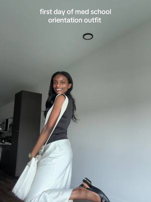 A young woman in a stylish outfit poses for a photo, angled to her side while holding a bag. She's smiling, wearing a black top, white pants, and black heels. The text overlay at the top says: first day of med school orientation outfit. The color scheme is simple, with the white wall and the neutral lighting adding to a clean, modern composition.