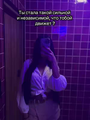 A person, likely a woman, is taking a selfie in a bathroom. The person is wearing a white button-down shirt. The background consists of square, purple tiles. The text overlayed is in the upper left corner.