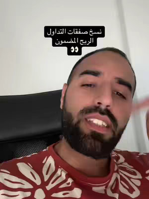 A selfie-style shot featuring a light-skinned man with a trimmed beard and short hair. He's wearing a red shirt with a white floral pattern. Behind him, a blurred background shows a plain wall. A black overlay contains Arabic text in a bold white font. He is speaking, with his mouth slightly open.