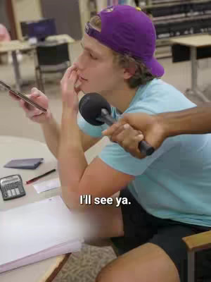 A young man in a purple cap and blue shirt is holding a phone and covering his mouth with his hand, as if in thought or surprise, while a black microphone is held towards him by another person. The scene appears to be in a casual indoor setting, possibly a library or office, with desks and other items in the blurred background.