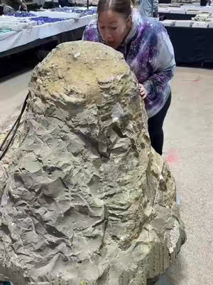 A woman leans in to sniff a large, irregularly shaped rock. The rock is a light tan color, with a rough, textured surface. The woman wears a colorful tie-dye jacket and black pants. The background suggests a large indoor space, possibly a market or convention hall, with tables and other people visible in the distance.