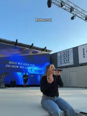 A woman sings into a microphone on an outdoor stage. Behind her is a large screen displaying lyrics, and in the background, a band is setting up. The sky is a clear blue.
