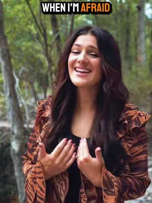 A woman with long dark hair smiles while outdoors against a blurred green background. The image has a warm color palette with autumn tones emphasized by her patterned top.