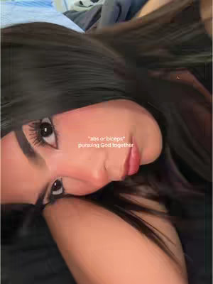 A close-up shot showing a woman's face tilted diagonally. Her dark hair partially obscures the upper part of the frame. Her eyes are open and gazing towards the viewer, and her face has full makeup with bright red lip color and blush. Text is overlaid on the bottom right of her face, and the background is a soft-focus blend of what appears to be a bed and other objects.