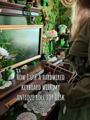 The cover image features a person holding a wired keyboard in front of an antique roll-top desk. The desk is adorned with various decorative items, including a framed painting, small figurines, and doilies. The background is filled with green plants and decorations, creating a cozy, maximalist aesthetic. The overall color scheme is dominated by greens and browns.