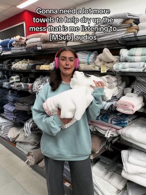 A woman wearing a light blue sweatshirt and gray sweatpants stands in a store aisle filled with folded towels, holding a stack of white towels and a pink-eared headphone. Above her, text overlays describe the scene.