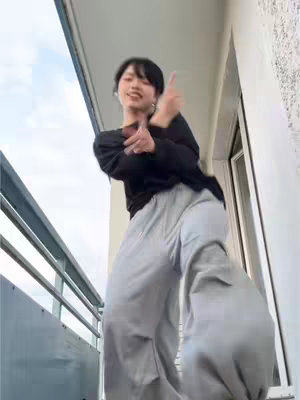 A young woman is filmed on a balcony, dancing with energetic poses while facing the camera. She wears a black long-sleeved top and loose gray pants. The background shows a clear sky and a white balcony railing and wall.