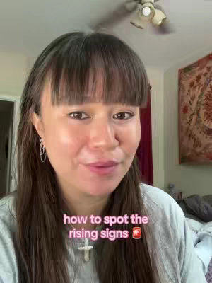 A woman with long brown hair and bangs is centered in the frame, smiling gently. Text overlaid on the image reads "how to spot the rising signs". The background shows a domestic interior with a ceiling fan and a tapestry.