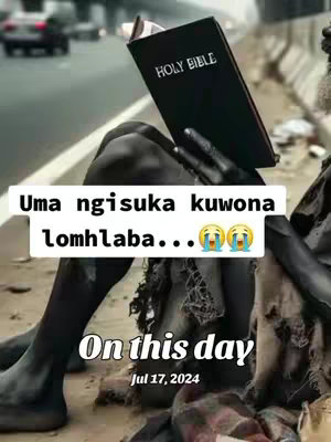 The image is a low-angle shot featuring a person sitting on the side of a road holding a bible. The individual seems to be of darker skin tone, with their skin covered in dark color. The person is looking down at the bible. The background is blurred, suggesting a busy road with cars. Overlayed on the image is a text overlay including text and a date.