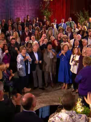 A stage is filled with a large group of people, mostly adults, some holding microphones as if they are singing. People in the foreground are applauding. The background features a brick wall and red curtains.