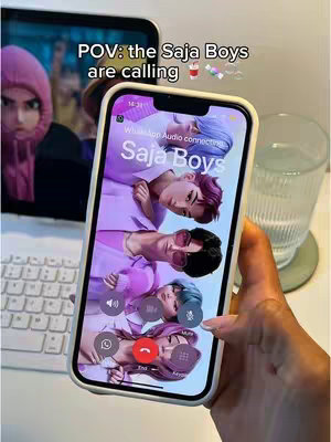 The image shows a hand holding a phone displaying a call screen with the names "Saja Boys" and their animated faces. The scene includes the phone, part of a laptop screen in the blurred background, and a glass of water.