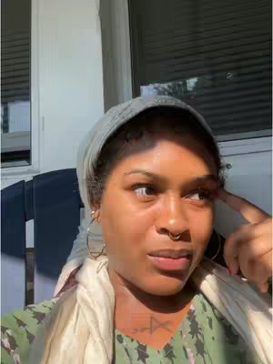 A woman with a neutral expression looks to the side, with a hand near her face, as if deep in thought. She wears a headscarf, large hoop earrings, and a necklace with a tattoo visible on her chest.