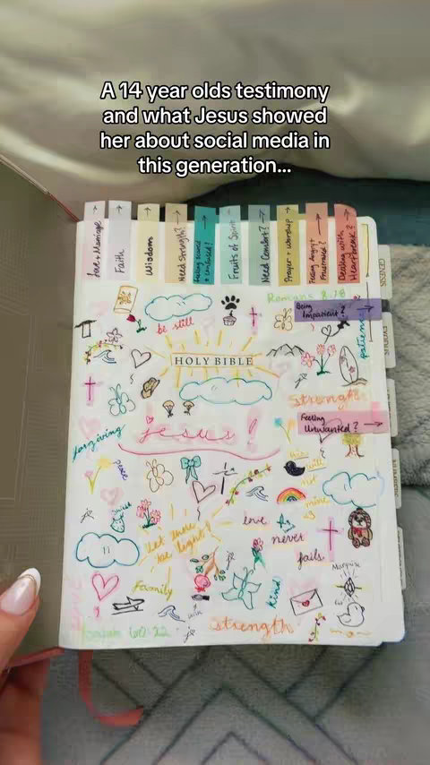 The image shows an open Bible with handwritten notes, drawings, and sticky notes scattered across the pages. The pages are filled with colorful annotations, including phrases like 'forgiving', 'Jesus!', and 'strength', alongside various doodles such as hearts, flowers, and clouds. Tabs with written labels such as "Faith" and "Wisdom" are visible along the top edge. A hand with painted nails is holding the book.