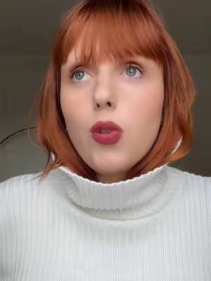 A close-up shot of a person with red hair and fair skin looking towards the camera with their mouth slightly open. They are wearing a white turtleneck. The background is out of focus.