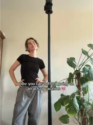 A woman stands with her hands on her hips, looking up towards a dark pole extending upwards. The scene is set indoors with a plant on the right and a beige wall in the background. Text overlay reads, 'pov: you can't afford going on an actual vacation.'