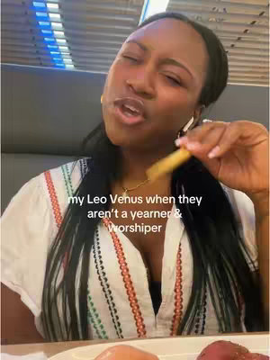 A dark-skinned woman with long, dark hair is holding a french fry in her right hand. She is wearing a white, patterned top with red and orange accents around the neckline. She appears to be mid-sentence, mouth open in an inquisitive expression. The background suggests she is in a restaurant.