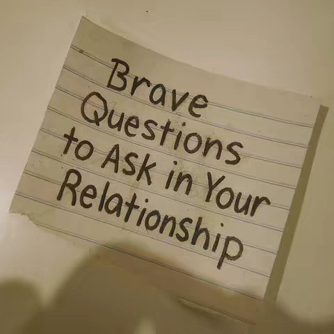 A photograph of a slightly crumpled piece of lined paper against a white vertical surface, perhaps a wall or table. The paper has handwritten text in a dark, imperfect font that reads: 'Brave Questions to Ask in Your Relationship'. The lighting is soft and diffused, creating gentle shadows to the bottom-right of the paper. The background provides negative space with little visual detail, thus making the text on the paper the main focus.