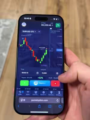 A close-up shot focuses on a person's hand holding a black smartphone. The phone's screen displays a trading app interface with a candlestick chart, representing financial data. The background seems to be a wooden surface, and the overall lighting is natural.