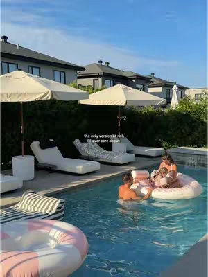 A photo captures a luxurious outdoor setting on a bright day. In the foreground, a swimming pool with clear blue water dominates the scene. Several people are in the pool, some sitting on pink and white inflatable rings. Behind them, a patio area features white lounge chairs and matching umbrellas, suggesting a relaxing area. A series of modern buildings with dark roofs and clean lines forms the backdrop, with a clear sky overhead adding to the serene and upscale atmosphere.