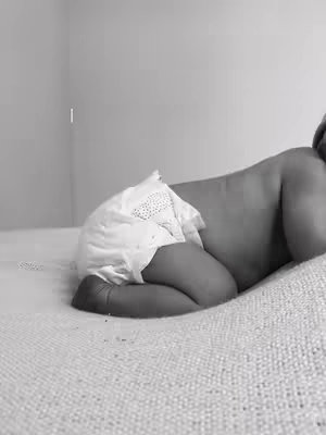 A grayscale image shows a baby lying prone on a textured blanket, with the baby's back and a portion of its side visible. The baby wears a diaper. Soft lighting and a minimalist background complete the scene.