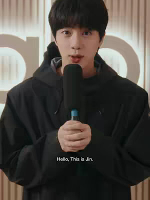 The image features a medium close-up of Jin, a man with black hair and a black hoodie, holding a microphone. The background includes a blurred view of the 'alo' logo and some lighter toned walls. The text "Hello, This is Jin." is overlaid on the bottom part of the image.
