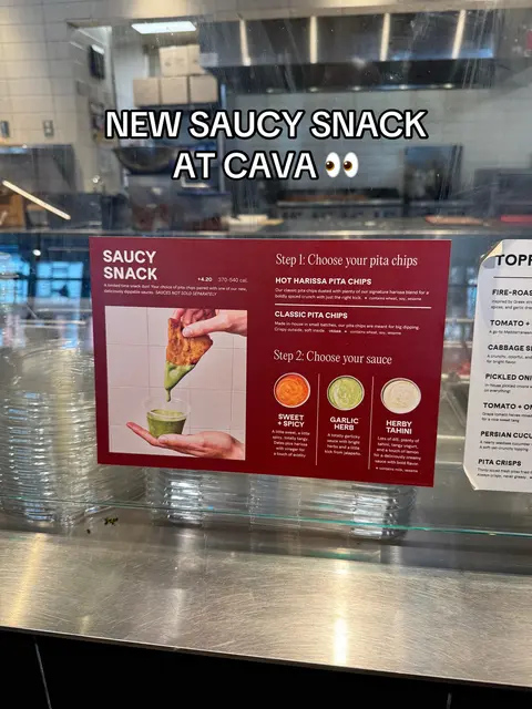 A view of a Cava restaurant display case. In the foreground, there's a sign about a new "Saucy Snack" with text overlays and graphic elements. The background reveals a stainless steel counter, kitchen equipment, and the restaurant's ceiling, featuring overhead lights.
