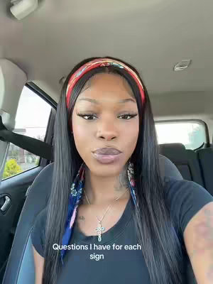 A woman is seated in a car, facing the camera, with a clear view of her face. She's wearing a headband with a red floral pattern. Her long black hair is parted down the middle. She's also wearing a necklace with a cross pendant. The background shows the car's interior and a glimpse of the outside surroundings.