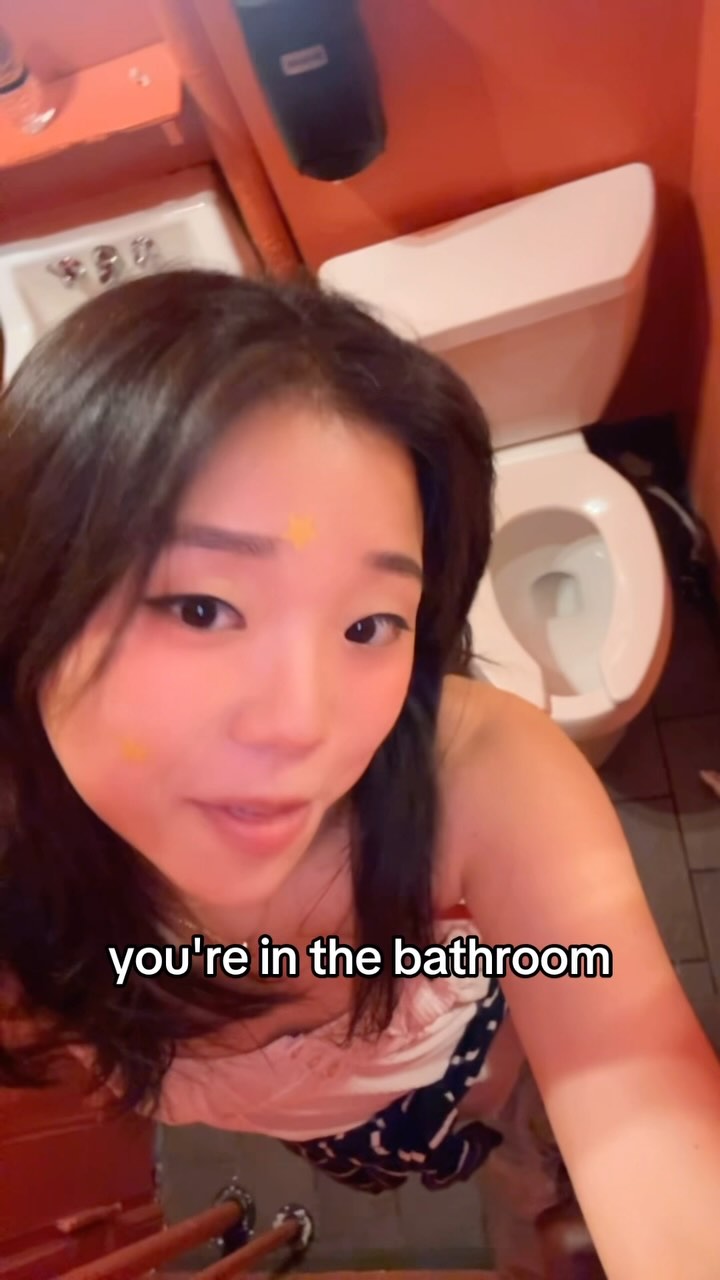 A selfie-style shot taken from above, featuring a young woman inside a bathroom. The woman is the central focus, framed against a backdrop of a toilet and orange painted walls. She has dark hair, and is looking at the camera with a small heart-shaped makeup sticker on her cheek. The text overlay in the image reads 'you're in the bathroom'.