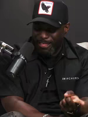 A close-up shot of a Black man wearing a black baseball cap, a black shirt, and a silver chain with a cross pendant. He is holding a microphone and speaking. The background is dark, and the lighting is dim.