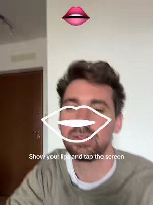 A man is in the foreground with a filter and overlaid text and graphics. Above the man's head is a graphic of red lips. Overlaying the man's face is a white outline of lips. The text 'Show your lips and tap the screen' is displayed on the bottom of the screen.