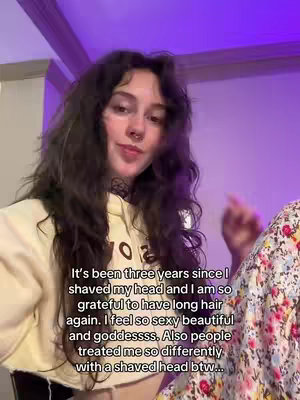 The cover image features a young woman with long, wavy dark hair in front of a purple-lit background. She is wearing a cream-colored hoodie and a black choker. Text overlays are present, stating the duration since she shaved her head and expressing gratitude for her hair's growth. The image is framed from the chest up, showcasing the woman's face and upper body as the central subjects.