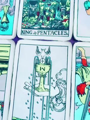 The image shows a close-up of several tarot cards arranged together. The central card displays a golden chalice held in a hand with water droplets falling from above. Surrounding cards are visible with text indicating the 'King of Pentacles.' The overall composition is artistic, emphasizing the details of the tarot imagery.