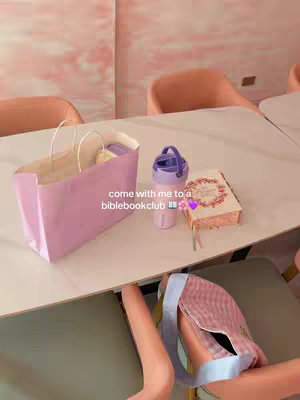 The image is taken from a high angle, showing a white table. On the table are several items: a light pink paper bag with white handles, a purple insulated water bottle, and a closed book with decorative pages. The table rests over a lower level with seats and a part of a person is visible.