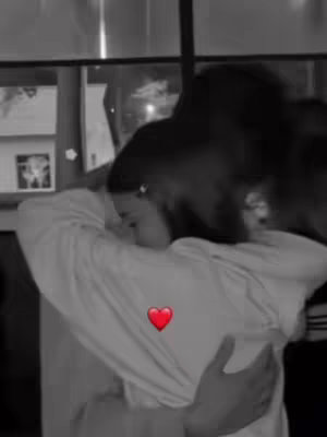 A black and white image shows two figures embracing. The person in the foreground, wearing a white long-sleeve shirt, has their back to the camera. The other person is hugging them from behind, their face obscured by shadows and hair. A red heart emoji is centered on the shirt.