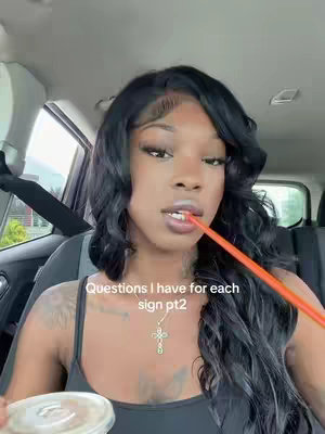 A woman with long, wavy black hair sits inside a car, holding a drink with a straw in her mouth. The scene is framed from the chest up, and the background shows the inside of the car with a glimpse of the outside world through the window. The woman is wearing a black top and a necklace. Text on the video overlay reads 'Questions I have for each sign pt2'.