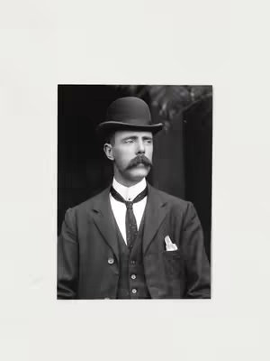 A black and white portrait of a man in a traditional suit and bowler hat. The man has a well-groomed mustache and is looking slightly off-center. There's a hint of a pocket square. The background is subtly blurred indicating a backdrop of a room or environment.